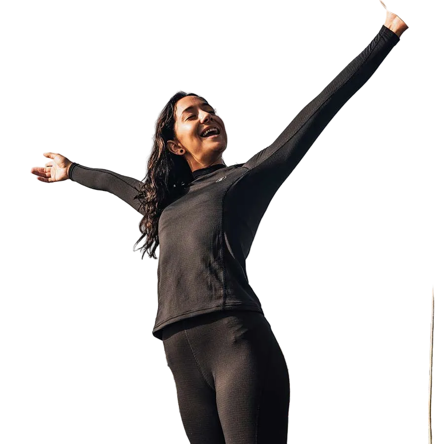 A person wearing a black, long-sleeved wet suit top standing with arms raised in a coastal location.